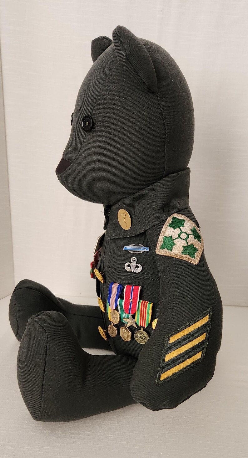 Op de afbeelding: Een donkergroene teddybeer met een militair uniform. De beer heeft een zwart knoopoog en een bruine neus. Het uniform bevat patches, medailles en strepen, wat duidt op een gedecoreerd lid. De beer zit.