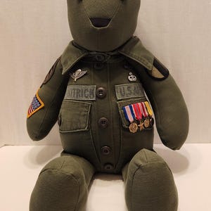 May include: A green teddy bear dressed in a military uniform with patches and medals. The bear is sitting on a white surface.
