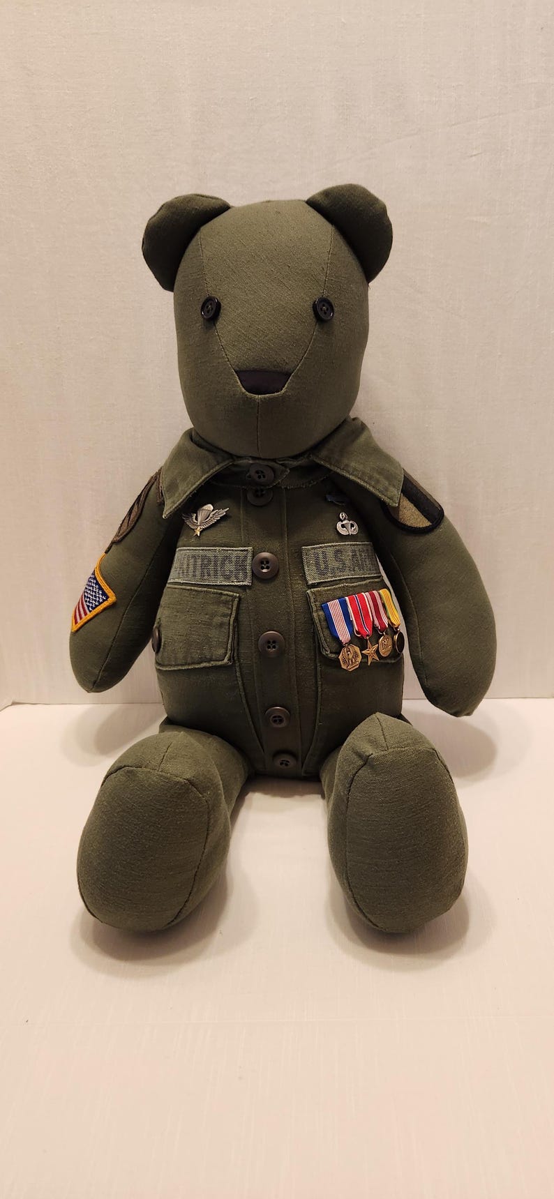 May include: A green teddy bear dressed in a military uniform with patches and medals. The bear is sitting on a white surface.