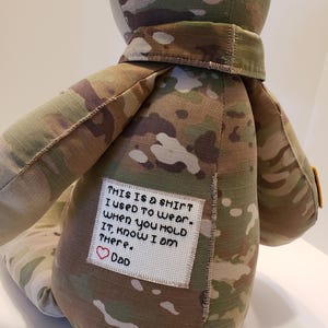 May include: A camouflage fabric teddy bear with a white patch sewn on the back. The patch has a message that reads "THIS IS A SHIRT I USED TO WEAR. WHEN YOU HOLD IT, KNOW I AM THERE. DAD" with a red heart symbol.