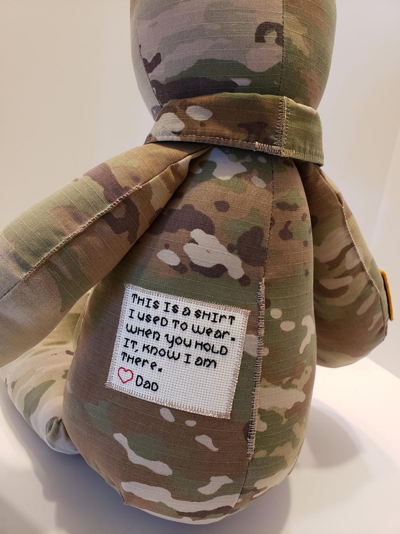 May include: A camouflage fabric teddy bear with a white patch sewn on the back. The patch has a message that reads "THIS IS A SHIRT I USED TO WEAR. WHEN YOU HOLD IT, KNOW I AM THERE. DAD" with a red heart symbol.
