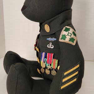 Op de afbeelding: Een donkergroene teddybeer gekleed in een militair uniform. Het uniform heeft patches, medailles en strepen. De beer heeft zwarte knoopogen en zit met uitgestrekte benen.