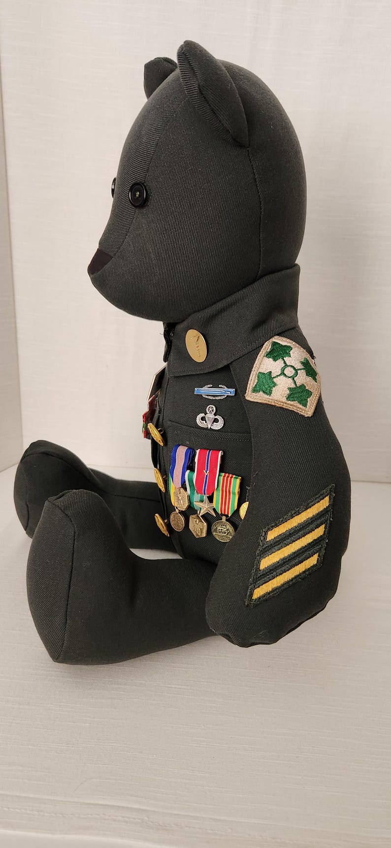 Op de afbeelding: Een donkergroene teddybeer gekleed in een militair uniform. Het uniform heeft patches, medailles en strepen. De beer heeft zwarte knoopogen en zit met uitgestrekte benen.