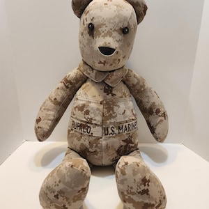 Op de afbeelding: Een teddybeer in een woestijn camouflagepatroon. De beer heeft zwarte knoopogen, een gestikte neus en mond. Op het uniform staan de namen "RUFALO" en "U.S. MARINES" geborduurd.