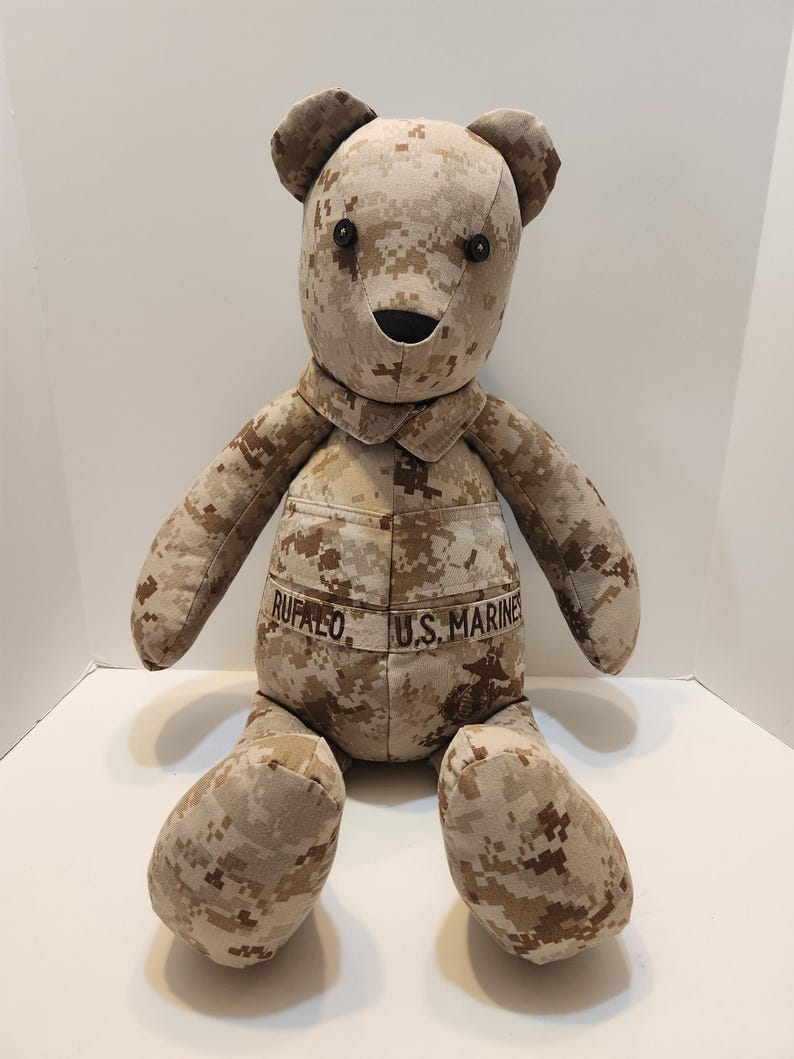 Op de afbeelding: Een teddybeer in een woestijn camouflagepatroon. De beer heeft zwarte knoopogen, een gestikte neus en mond. Op het uniform staan de namen "RUFALO" en "U.S. MARINES" geborduurd.