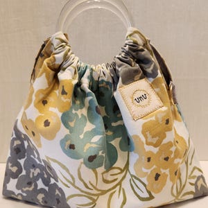 May include: A floral print handbag with a clear handle. The bag features a white background with yellow, blue, and gray flower patterns. The text "The Blooming Bag" and "VKUDESIGNS" are visible.