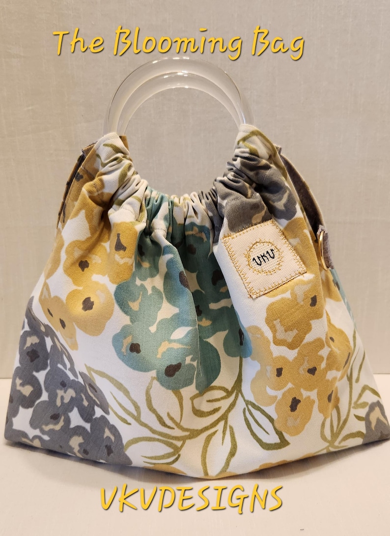 May include: A floral print handbag with a clear handle. The bag features a white background with yellow, blue, and gray flower patterns. The text "The Blooming Bag" and "VKUDESIGNS" are visible.