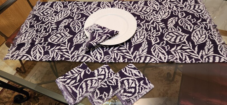 May include: A set of purple and white table linens with a leaf pattern. The set includes a table runner and four placemats.
