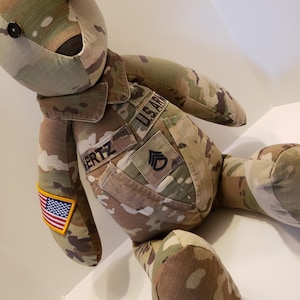 May include: A camouflage fabric teddy bear wearing a military uniform with the text "US ARMY" and "VERTZ" on the uniform. The bear has a US flag patch on its arm and a rank insignia on its chest.