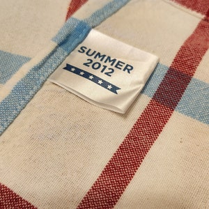 K&ouml;nnte beinhalten: Ein wei&szlig;er Stoff mit rotem und blauem Karomuster. Ein wei&szlig;es Etikett mit dem Text "SUMMER 2012" und vier Sternen ist auf den Stoff gen&auml;ht.