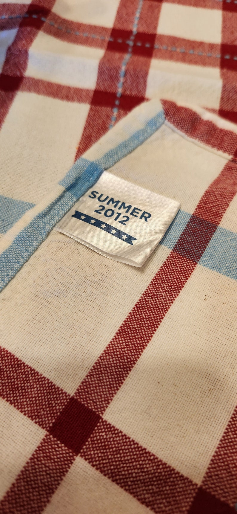 K&ouml;nnte beinhalten: Ein wei&szlig;er Stoff mit rotem und blauem Karomuster. Ein wei&szlig;es Etikett mit dem Text "SUMMER 2012" und vier Sternen ist auf den Stoff gen&auml;ht.