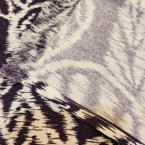 May include: A close-up of a white and purple fabric with a fringed edge. The fabric has a repeating pattern of abstract shapes.