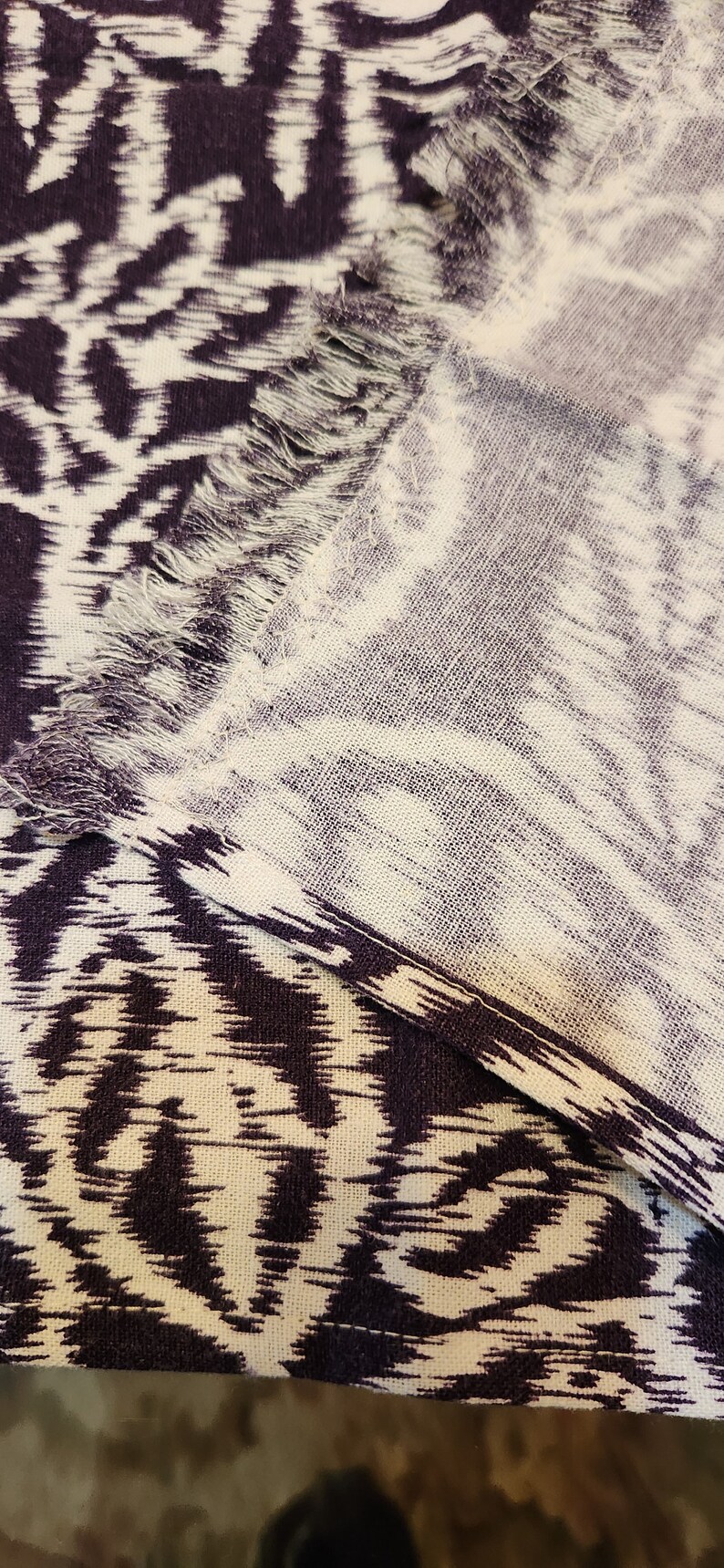 May include: A close-up of a white and purple fabric with a fringed edge. The fabric has a repeating pattern of abstract shapes.