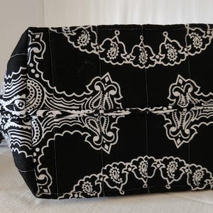 May include: A black and white quilted zippered pouch featuring a repeating paisley pattern. The rectangular bag has vertical stitching lines across the fabric and includes a small matching fabric pull tab attached to the side zipper closure.