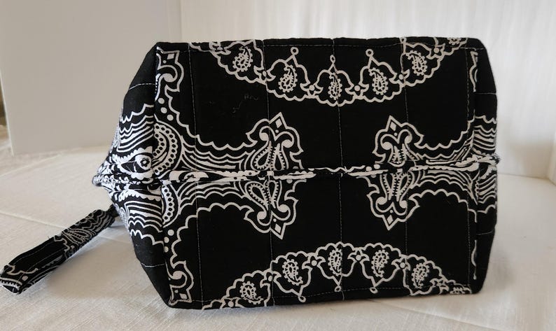 May include: A black and white quilted zippered pouch featuring a repeating paisley pattern. The rectangular bag has vertical stitching lines across the fabric and includes a small matching fabric pull tab attached to the side zipper closure.