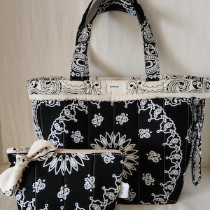 May include: A black and white quilted tote bag and a matching zippered pouch, both featuring a traditional paisley bandana print. The tote has two handles and a small white label with the text UKU. The pouch includes a decorative fabric knot.
