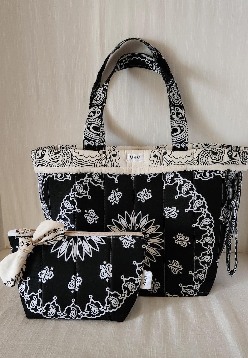 May include: A black and white quilted tote bag and a matching zippered pouch, both featuring a traditional paisley bandana print. The tote has two handles and a small white label with the text UKU. The pouch includes a decorative fabric knot.