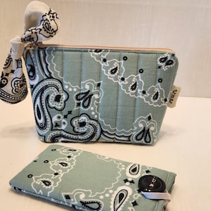 May include: A sage green quilted zippered pouch with a black and white paisley bandana print and a matching fabric bow attached to the zipper pull, accompanied by a rectangular sage green paisley print case featuring a black button closure.
