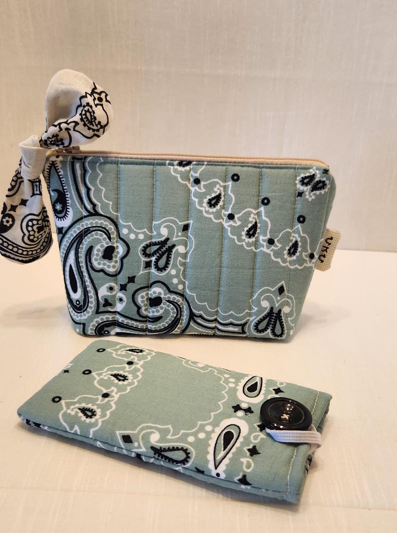 May include: A sage green quilted zippered pouch with a black and white paisley bandana print and a matching fabric bow attached to the zipper pull, accompanied by a rectangular sage green paisley print case featuring a black button closure.