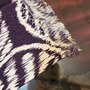 May include: A close-up of a purple and white patterned fabric with a fringed edge. The fabric has a subtle ikat design.