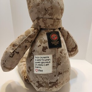 Op de afbeelding: Een teddybeer gemaakt van camouflage stof, met een sjaal en patches. E&eacute;n patch leest "AMERICAN APPAREL INC." en een andere heeft geborduurde tekst: "This is pants I used to wear. When you hold it, know I am there. ❤️ Max."