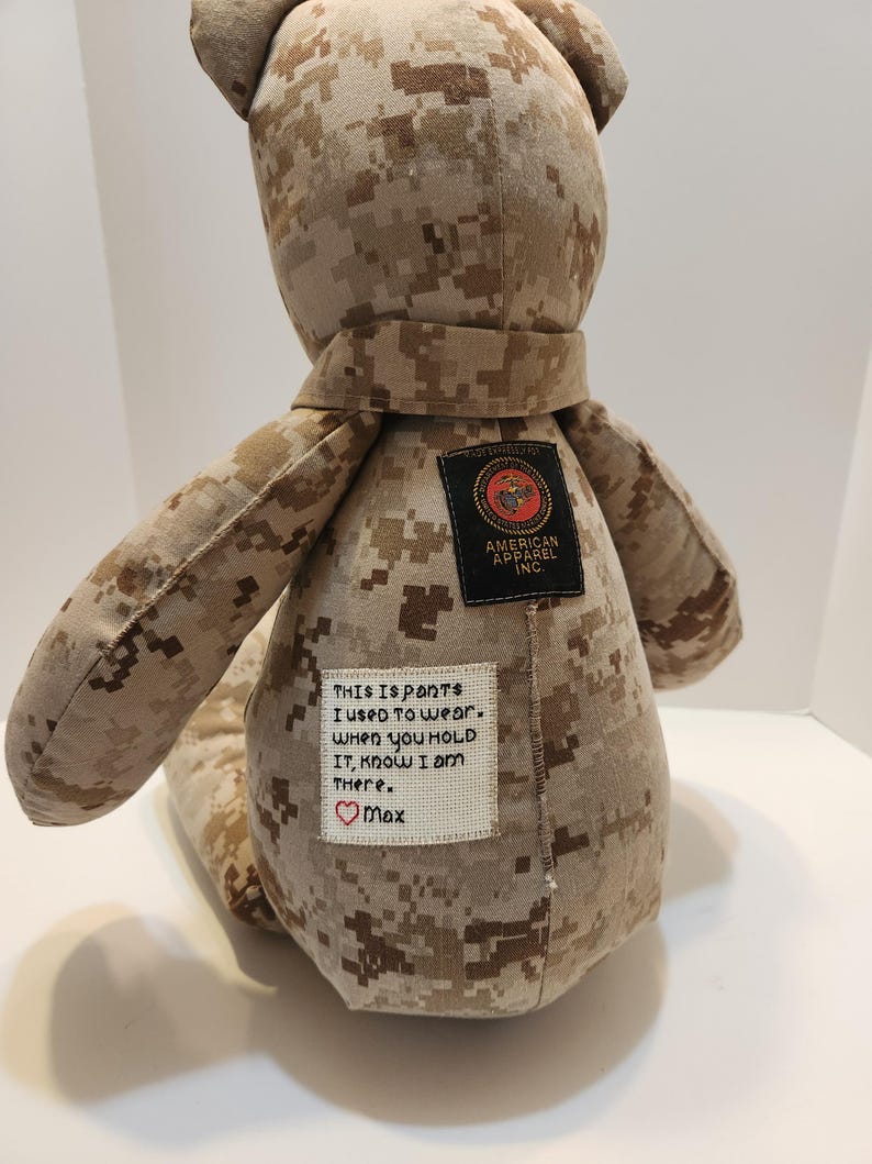 Op de afbeelding: Een teddybeer gemaakt van camouflage stof, met een sjaal en patches. E&eacute;n patch leest "AMERICAN APPAREL INC." en een andere heeft geborduurde tekst: "This is pants I used to wear. When you hold it, know I am there. ❤️ Max."