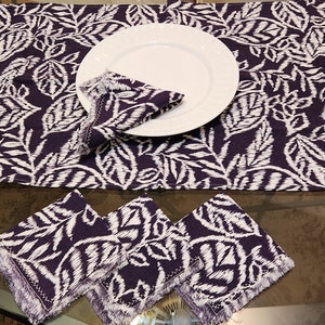 May include: A set of purple and white table linens with a leaf pattern. The set includes a table runner and three napkins. The napkins have fringed edges.