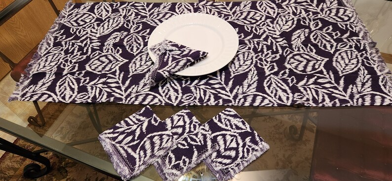 May include: A set of purple and white table linens with a leaf pattern. The set includes a table runner and three napkins. The napkins have fringed edges.