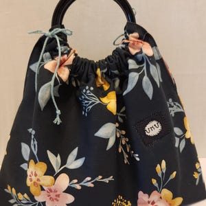 May include: Black fabric bag with a floral pattern in yellow, pink, and blue. The bag has a drawstring closure and a black handle. A small square patch with the text "UKU" is sewn on the side.
