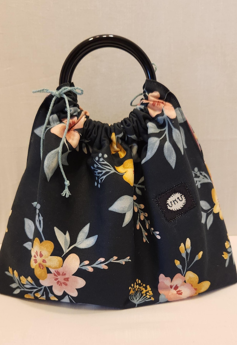 May include: Black fabric bag with a floral pattern in yellow, pink, and blue. The bag has a drawstring closure and a black handle. A small square patch with the text "UKU" is sewn on the side.