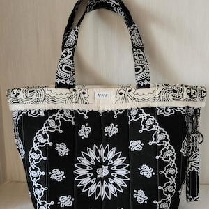 May include: A black and white quilted tote bag featuring a repeating paisley bandana pattern. The bag has two matching fabric handles, a top border with a cream-colored trim, and a small white rectangular label with the text UKU centered on top.