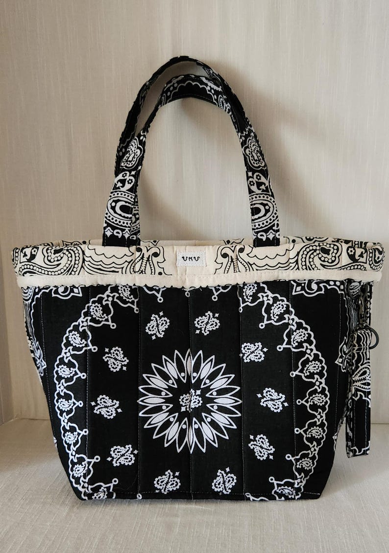 May include: A black and white quilted tote bag featuring a repeating paisley bandana pattern. The bag has two matching fabric handles, a top border with a cream-colored trim, and a small white rectangular label with the text UKU centered on top.