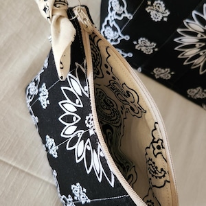 May include: A black and white zippered pouch featuring a paisley bandana pattern with a cream-colored fabric tie attached to the zipper pull. The interior lining is cream-colored with a matching black paisley print. A larger matching bag is visible behind.
