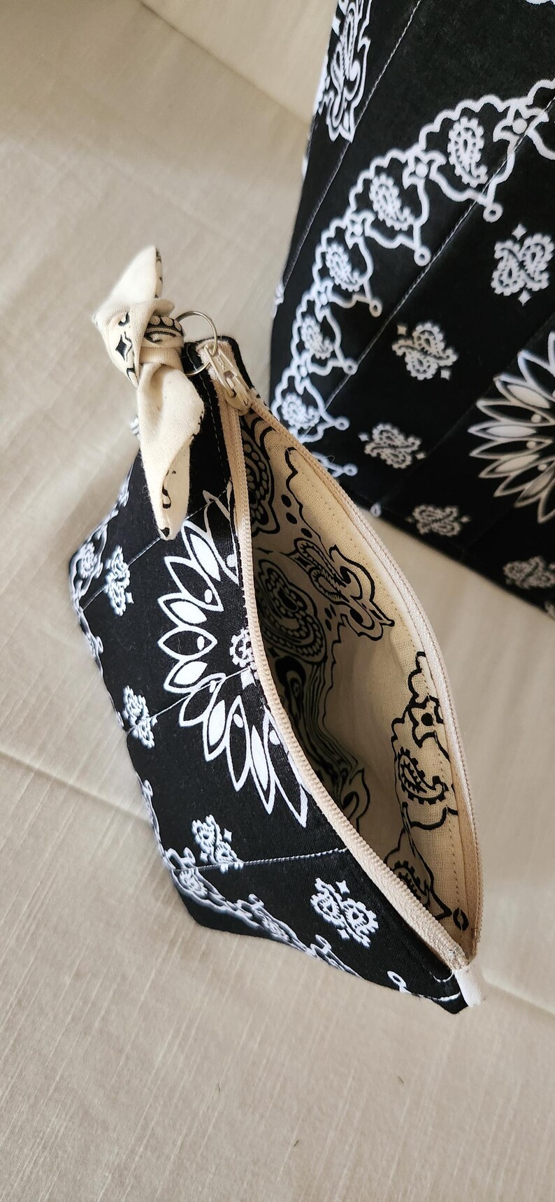 May include: A black and white zippered pouch featuring a paisley bandana pattern with a cream-colored fabric tie attached to the zipper pull. The interior lining is cream-colored with a matching black paisley print. A larger matching bag is visible behind.