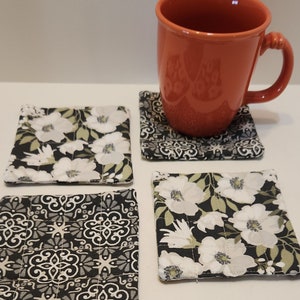 May include: Four fabric coasters with different patterns. Two coasters feature a black background with white flowers and green leaves. The other two coasters feature a black background with a white and gray geometric pattern.