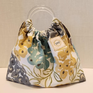 May include: A floral print handbag with clear acrylic handles. The bag features a white background with yellow, teal, and gray floral patterns. A small tag with the text "UKU" is sewn on the side. The bag is gathered at the top.