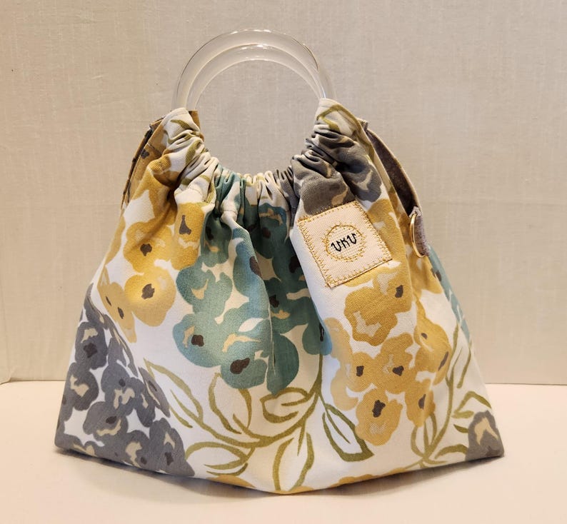 May include: A floral print handbag with clear acrylic handles. The bag features a white background with yellow, teal, and gray floral patterns. A small tag with the text "UKU" is sewn on the side. The bag is gathered at the top.