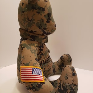 Op de afbeelding: Een teddybeer gemaakt van camouflage stof. De beer heeft een zwart knoopoog en een kleine Amerikaanse vlag patch op zijn arm. De naam "HARREN" is op de borst van de beer geborduurd.