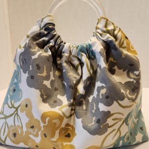 May include: A fabric handbag with a clear, circular handle. The bag features a floral pattern in shades of gray, yellow, and teal on a white background. The bag is gathered at the top, creating a rounded shape.