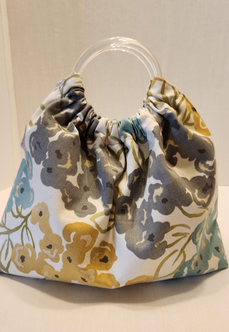 May include: A fabric handbag with a clear, circular handle. The bag features a floral pattern in shades of gray, yellow, and teal on a white background. The bag is gathered at the top, creating a rounded shape.