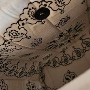 May include: The interior of a fabric bag featuring a white background with a black paisley bandana print pattern. The bag is constructed with quilted stitching and includes a single black circular snap button closure located at the top center.