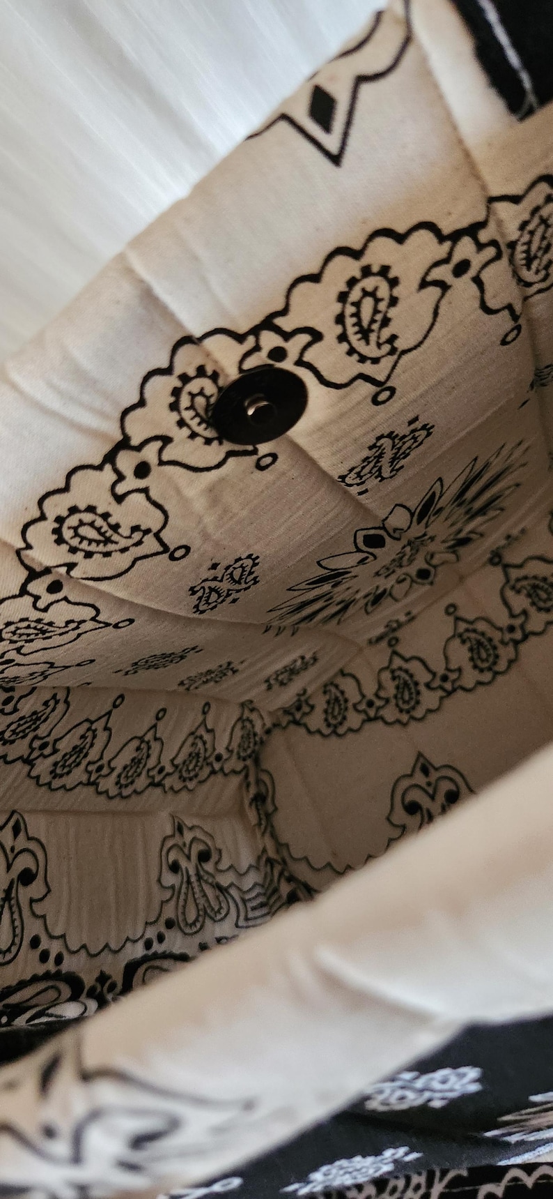 May include: The interior of a fabric bag featuring a white background with a black paisley bandana print pattern. The bag is constructed with quilted stitching and includes a single black circular snap button closure located at the top center.