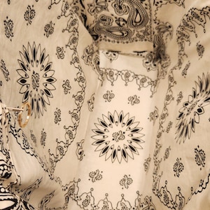 May include: A fabric tote bag with an open top, featuring an off-white base color and a repeating black bandana-style paisley and floral print. The interior includes a side pocket with a zipper closure and a small gold-toned metal ring attachment.