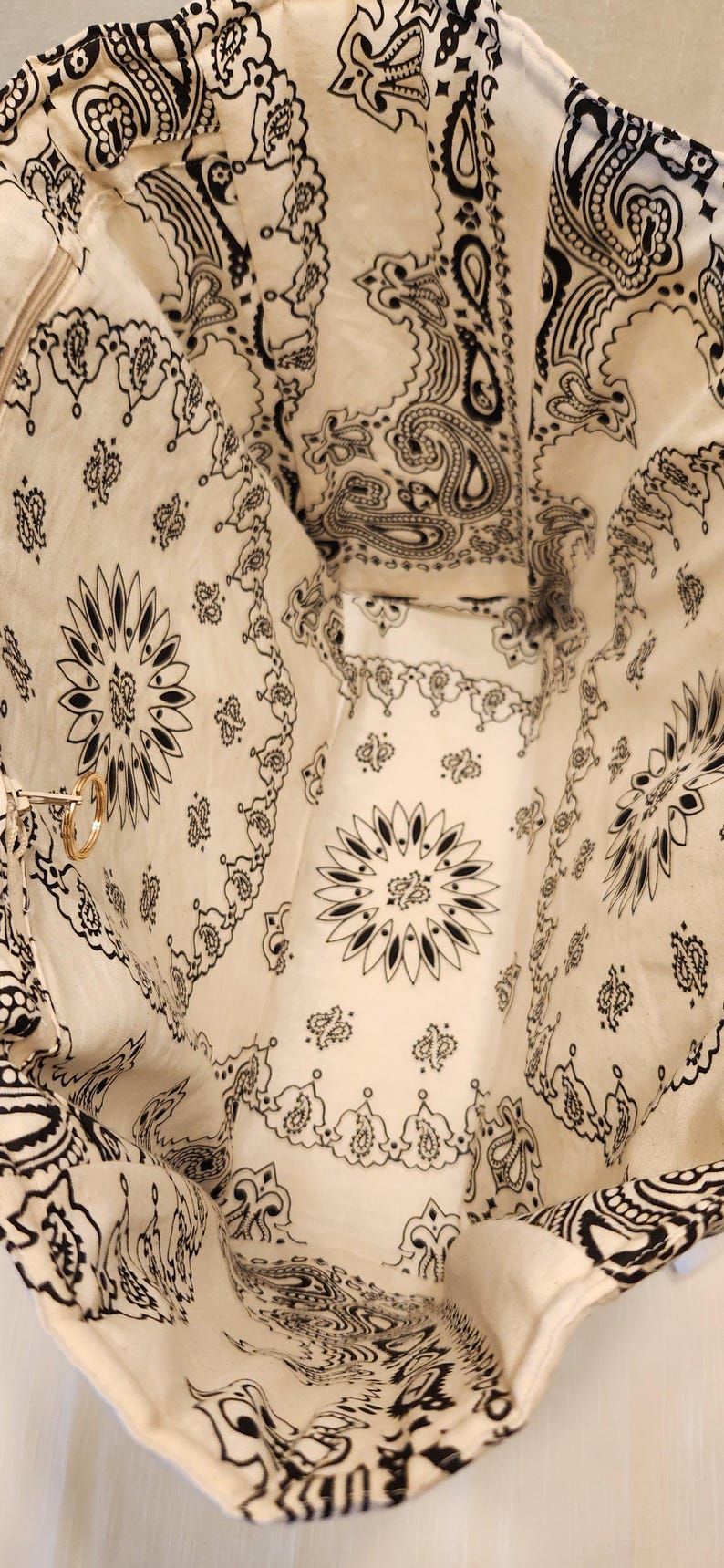 May include: A fabric tote bag with an open top, featuring an off-white base color and a repeating black bandana-style paisley and floral print. The interior includes a side pocket with a zipper closure and a small gold-toned metal ring attachment.