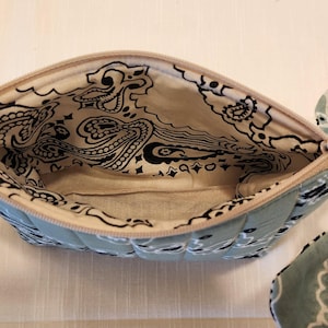 May include: A quilted zippered pouch featuring a light blue and white paisley bandana print. The interior is lined with a matching cream-colored fabric displaying a black paisley pattern. A matching fabric wrist strap is attached to the zipper pull.