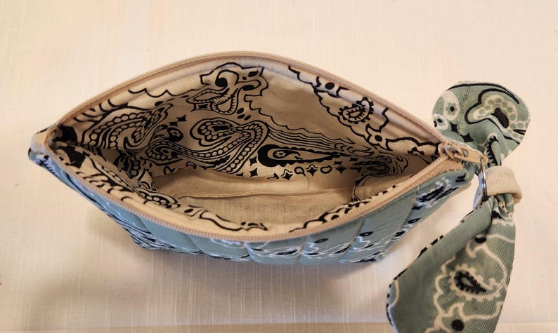 May include: A quilted zippered pouch featuring a light blue and white paisley bandana print. The interior is lined with a matching cream-colored fabric displaying a black paisley pattern. A matching fabric wrist strap is attached to the zipper pull.