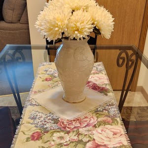 May include: A cream-colored vase with floral relief holds a bouquet of large, light yellow artificial flowers. The vase sits on a floral patterned table runner with a cream-colored center, placed on a glass table.