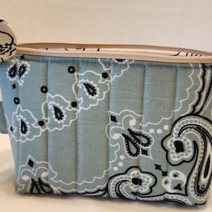 May include: A quilted zippered pouch featuring a light sage green fabric with a black and white paisley bandana pattern. The rectangular bag has vertical stitching lines across the front and a beige zipper closure along the top edge.