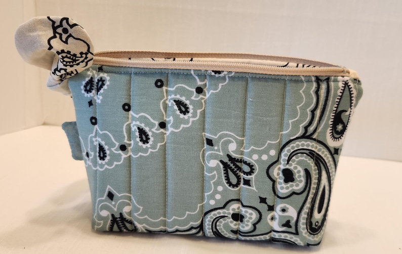 May include: A quilted zippered pouch featuring a light sage green fabric with a black and white paisley bandana pattern. The rectangular bag has vertical stitching lines across the front and a beige zipper closure along the top edge.