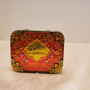 May include: Rectangular Whitman's Cloisonne tin with a red background and gold accents. The tin features floral and bird designs, with the words "Whitman's Cloisonne" in gold. The tin also says "Assorted Chocolates".